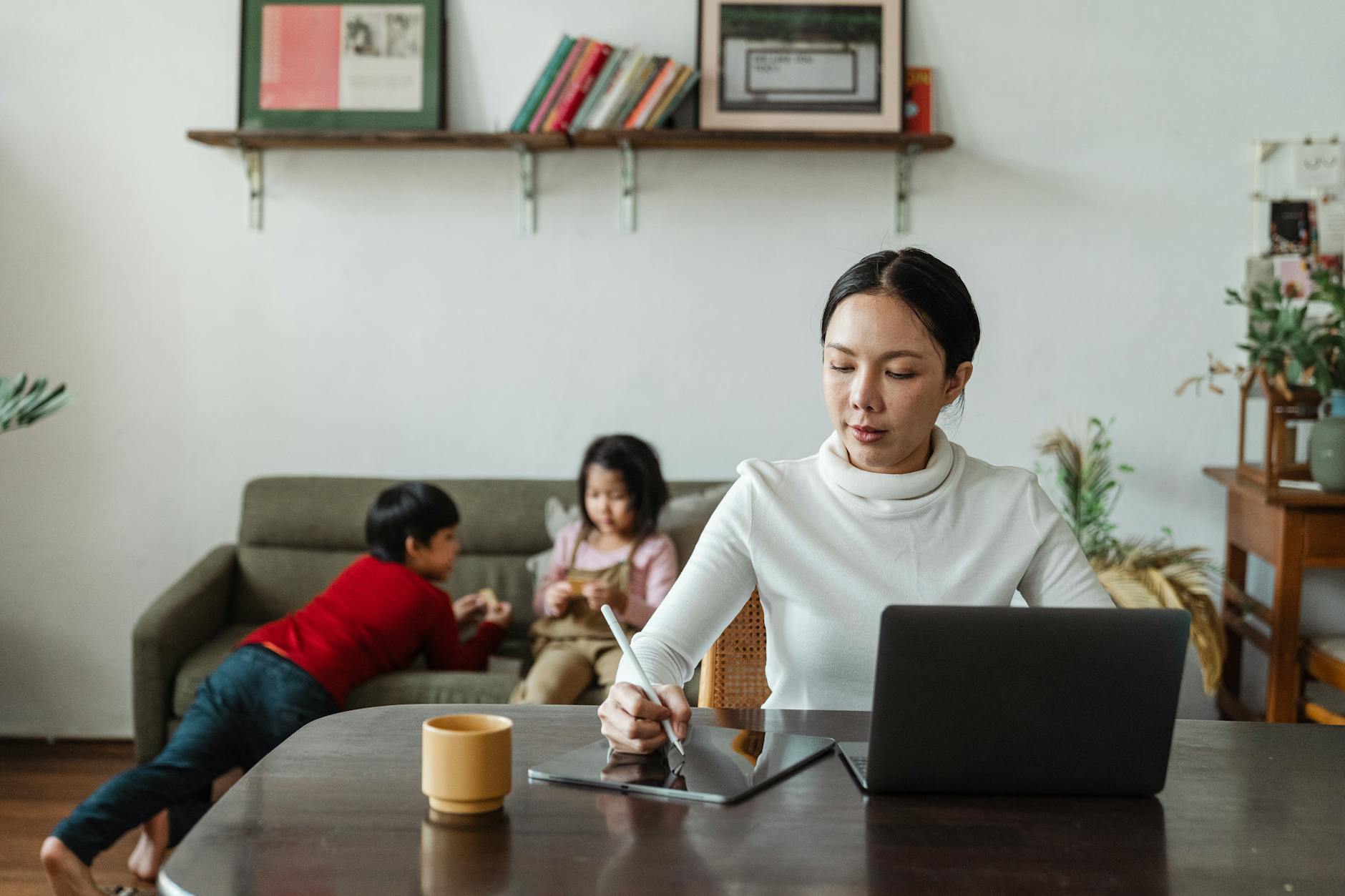 focused young asian mother working remotely while children playing on sofa